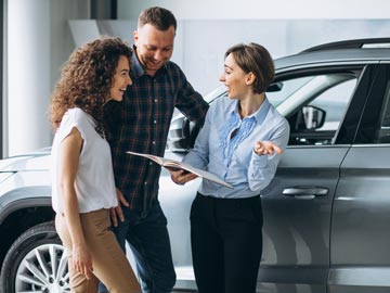 Ein Autoverkäufer bei Autoankauf Steinfurt bespricht Details mit einem Paar und hält dabei ein Broschüre in der Hand, in einem modernen Autohaus, mit Fahrzeugen im Hintergrund.